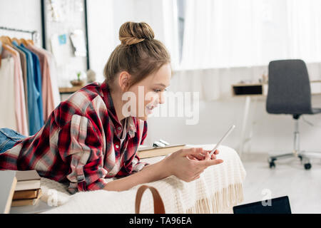Smiling teenager in checkered shirt lying on bed and using smartphone Stock Photo