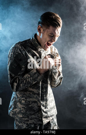 emotional man in military uniform screaming on black with smoke Stock Photo