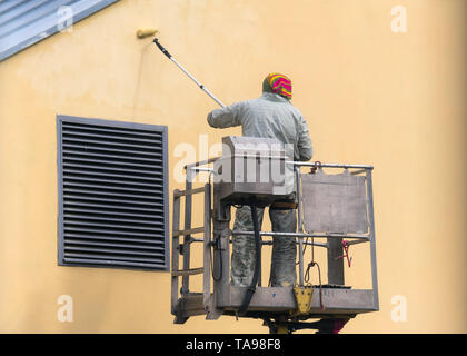 Man on a lifting platform painting the building wall with a roller exterior outdoors. Worker on a ladder manually painting yellow wall on construction Stock Photo