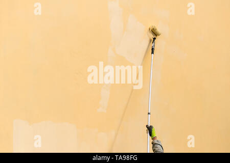 Painter paints building exterior wall with a roller. Roller with long stick manually painting building with yellow paint