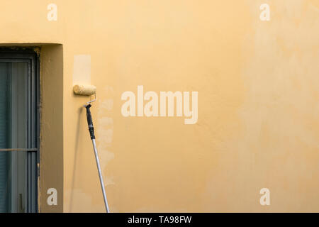 Painter paints building exterior wall with a roller. Roller with long stick manually painting building with yellow paint