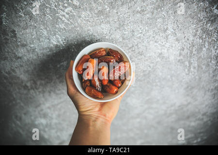 Dried whole dates in the white bowl holding in hand taken from the top on a gray and white background. Stock Photo