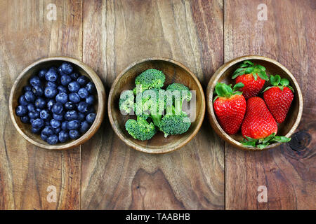 Three small wooden bowls are filled with healthy fresh fruits and vegetables and sitting on a rustic wooden plank background. Stock Photo