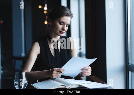 Accountant woman checking paperwork while sitting near window in creative office or cafe. Confident young woman working with documents.