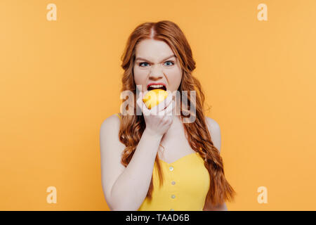 redhead girl looking at camera, making facial expression and biting lemon isolated on yellow Stock Photo