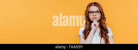 panoramic shot of beautiful pensive redhead girl in glasses propping chin isolated on yellow with copy space Stock Photo