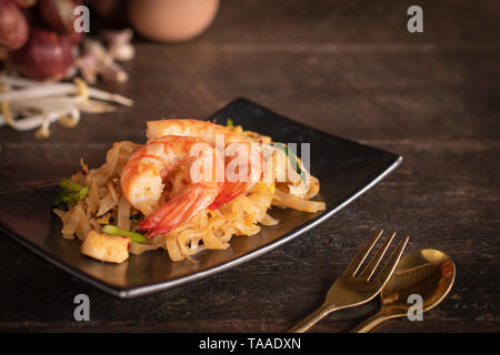 Thai fried noodles or pad thai with shrimp on black plate placed on the wood table there are fork and spoon place in front of and eggs, garlic, bean s Stock Photo