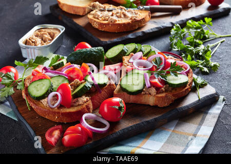 toasts with Cod Liver cream, topped with sliced cucumber, tomatoes and red onion rings on a cutting board with ingredients on a kitchen table, horizon Stock Photo