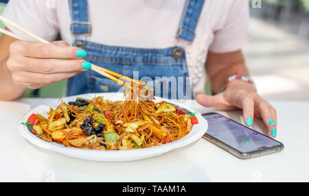 Woman eating chinese noodle with chicken and messaging on smartphone closeup