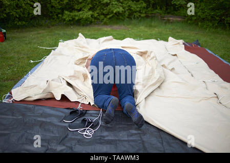 Funny image of a woman with her head inside a tent and her behind sticking out as she attempts to set up the tent outside on a camping ground