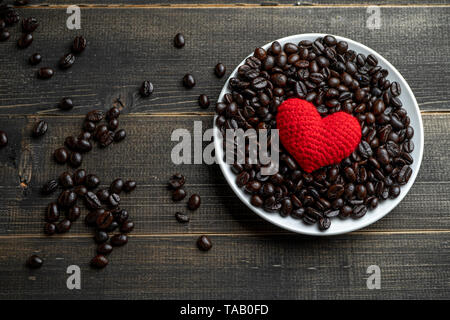 red heart on a lot of roasted coffee beans background on white plate on wooden table. Strong black espresso, Grains of coffee background, texture Stock Photo