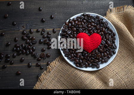 red heart on a lot of roasted coffee beans background on white plate on wooden table. Strong black espresso, Grains of coffee background, texture Stock Photo