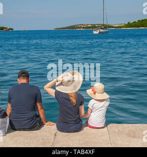 rear view of the family sitting on a stone pier on a summer day. The family admires the beautiful sea view from the shore. Stock Photo