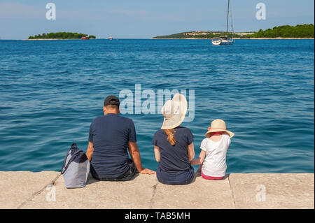 rear view of the family sitting on a stone pier on a summer day. The family admires the beautiful sea view from the shore. Stock Photo
