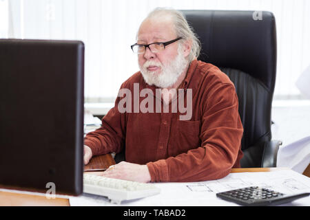 An elderly man in glasses sits at a table and works on computer in an office Stock Photo