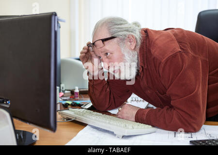 Senior man male sitting at desk wearing shirt, having trouble or problems using a computer, looking at monitor Stock Photo