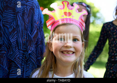 Cute little blonde girl with big blue eyes wearing a crown and happily smiling into camera during a birthday party in the park in summer sunshine Stock Photo