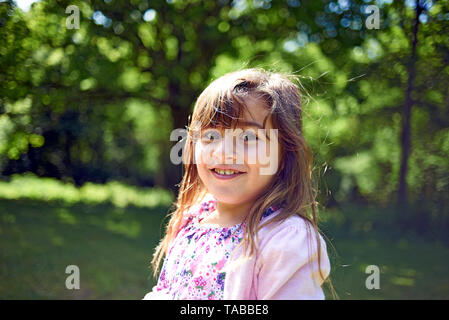 Cute girl with long brown hair looking at camera and smiling in spring sunshine in the park Stock Photo