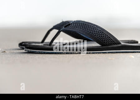 Selective-focus, low-angle view of black sandals on wet beach sand. Stock Photo