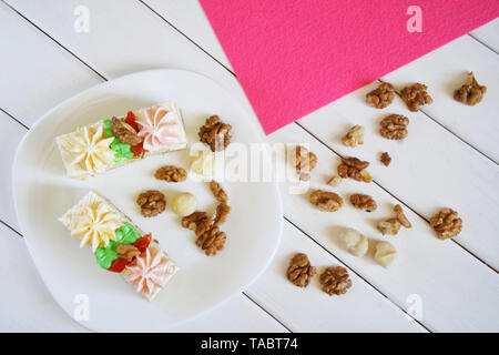 Two biscuit cakes with cream decorations are lying in a plate, next to sprinkled macadamia and walnuts on a white and pink background. Wooden table an Stock Photo