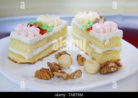 Sponge cakes with cream flowers, soaked in brandy, lie in a plate next to scattered macadamia nuts and walnuts on a light background. Wooden table. Cl Stock Photo