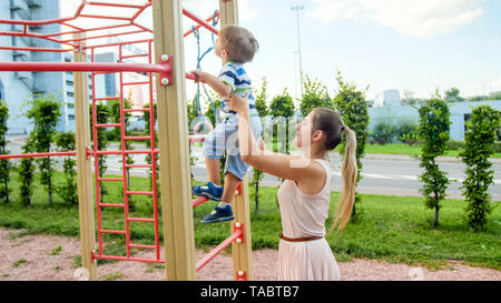 Portriat of young mother supporting and holding her 3 years old child son on metal ladder at children playground in park Stock Photo