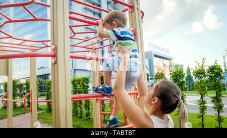 Portriat of young mother supporting and holding her 3 years old child son on metal ladder at children playground in park Stock Photo