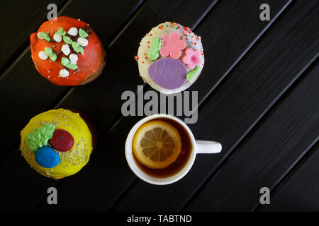 Three delicious, multi-colored cupcakes - Easter cakes on a black wooden surface and a cup of tea with a slice of lemon. Country breakfast. Homemade.  Stock Photo