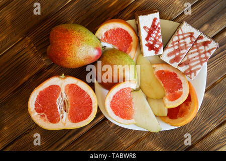 Three biscuit cakes, slices of grapefruit and pears lie in a white plate on a wooden table made of pine boards. Buffet in authentic natural hotel. Sum Stock Photo