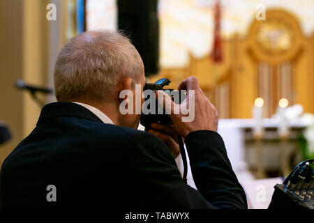 A gray-haired man in a suit takes pictures during church ceremonies. Shot from the back, blurry altar in the background. Stock Photo