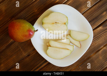 Sliced red pear lies in a white plate on a natural wooden background of pine boards. Summer still life. Daylight. Stock Photo