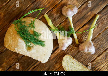 Garlic, dill sprigs and a white round bread bun are lying on a wooden table made of pine boards. Summer still life. Daylight. Stock Photo