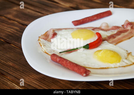 Small sausages, scrambled eggs with bacon and red chilli are in a white plate on a natural wooden surface of pine boards. Daylight. Stock Photo