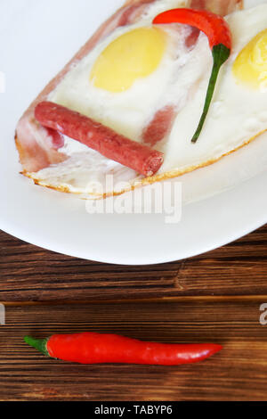 Red chili peppers, small sausages and bacon and eggs are in a white plate on a natural wooden surface of pine boards. Daylight. Vertical image Stock Photo