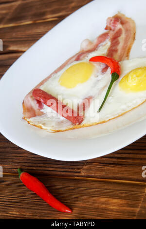 Fried eggs with bacon, red chili pepper and small sausages lie in a white plate on a natural wooden surface of pine boards. Daylight. Vertical image. Stock Photo