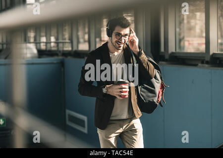 Smiling young man with headphones, cell phone and takeaway coffee walking at the station Stock Photo