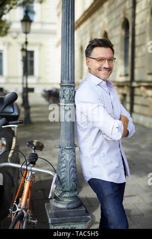 Portrait of smiling businessman with bicycle leaning against lamp post in the city Stock Photo