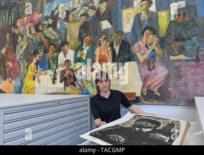 20 May 2019, Brandenburg, Beeskow: Florentine Nadolny, head of the art archive, is standing in front of rolling shelves in the new depot. In the background is the large-format painting by Christian Heinze 'Aus dem Leben Ernst Thälmanns' from 1982, which was commissioned by the Free German Trade Union Federation (FDGB). 30 years after the fall of communism, GDR art is finding interest again, especially among those who did not experience this state for themselves. The Beeskow Art Archive wants to make a contribution to this with its new open depot. It has around 23,000 GDR commissioned works of Stock Photo