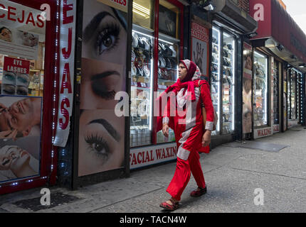A Muslim woman in a brightly colored dress and hijab walking past an eyebrow salon under the elevated subway on Roosevelt Ave. in Jackson Heights, NYC Stock Photo
