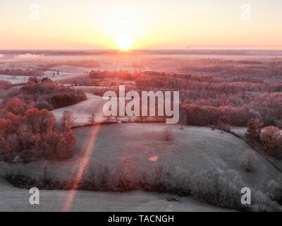 Aerial view of a frosty misty landscape at sunrise with low rolling hills, woodland trees and a fiery sun rising over the horizon Stock Photo