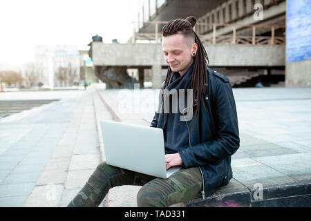 Young fashionable caucasian man with dreadlocks dressed in leather jacket sitting on the stairs using laptop. Remote works IT specialist Stock Photo