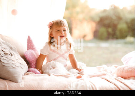 Laughing baby girl 1-2 year old wearing princess dress sitting in bed with pillow. Birthday party. Childhood. Stock Photo