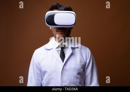 Studio shot of young handsome Hispanic man doctor using virtual reality headset against brown background Stock Photo