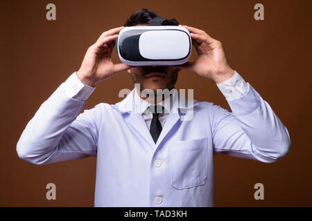 Studio shot of young handsome Hispanic man doctor using virtual reality headset against brown background Stock Photo