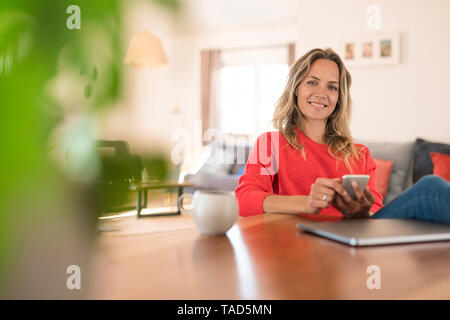 Portrait of smiling woman with laptop and cell phone on dining table at home Stock Photo