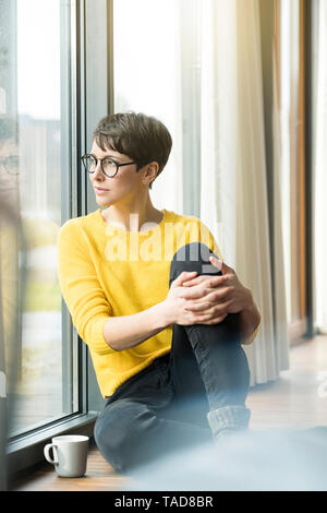 Portrait of woman with cup of coffee sitting on the floor at home looking out of window Stock Photo