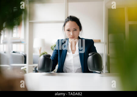 Portrait of young businesswoman sitting at desk wearing boxing gloves Stock Photo