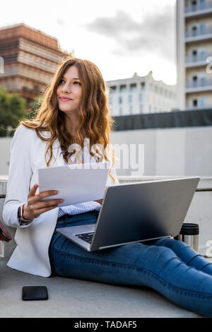 Young businesswoman sitting on stairs in the city, working with laptop, holding document Stock Photo