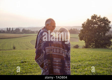 Happy senior couple standing on a meadow at sunset sharing blanket Stock Photo
