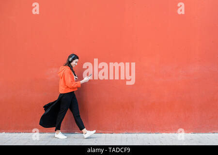 Young dancer walking and using the phone and headphones in front of a red wall